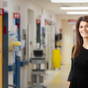 A healthcare professional wearing a stethoscope stands in a hospital hallway, smiling at the camera, with treatment rooms and medical equipment visible in the background.