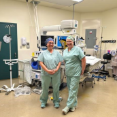 Two people stand side-by-side in an operating room wearing hospital scrubs.