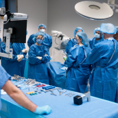 A group of people wearing blue surgical gowns stand around a hospital bed in a surgery room.