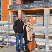 Image of a man (left) and woman (right) standing in front of a building and a construction fence.