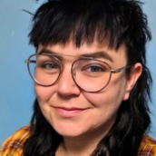 Close up image of a woman with long black hair and bangs. Wearing glasses and a yellow plaid shirt. There is a blue background behind her.