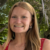 Close up image of a woman with long dirty blonde hair. Smiling largely. Standing outside with greenery behind her. She is wearing a short sleeve red polka dot top.