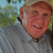 A man wearing a beige baseball cap and a gray collared shirt smiles gently at the camera, with soft greenery and blue sky in the background.