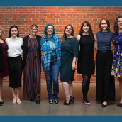 Image of 7 women standing in front of a brick wall smiling at the camera, professionally dressed.