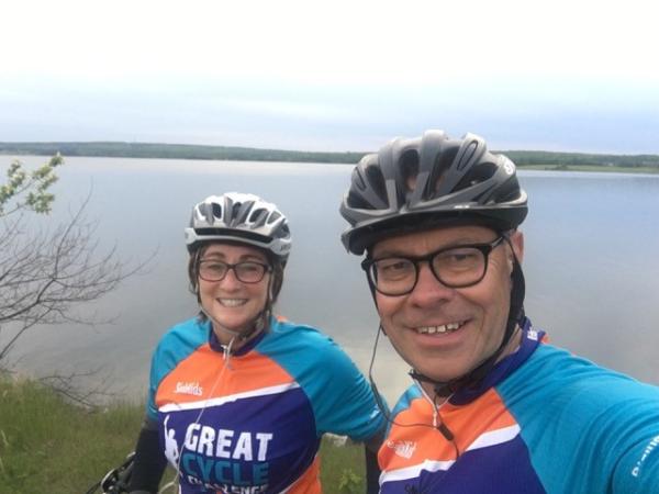 A woman wears glasses, a bike helmet and bike racing shirt, as does the man standing next to her. They are smiling at the camera, taking a selfie.