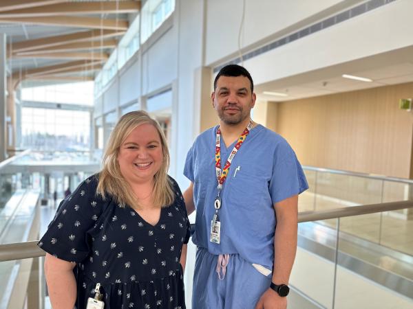 A woman stands with a man, facing the camera. She has long blonde hair, a black and white shirt and he is a doctor with medical scrubs, blue, and a lanyard.