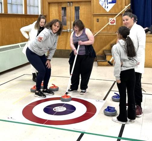 A group of individuals is engaged in a game of curling, showcasing teamwork and strategy. They are dressed in appropriate clothing for the sport and are positioned on a clean indoor surface. The scene includes both men and women actively participating, emphasizing the social aspect of this activity. 