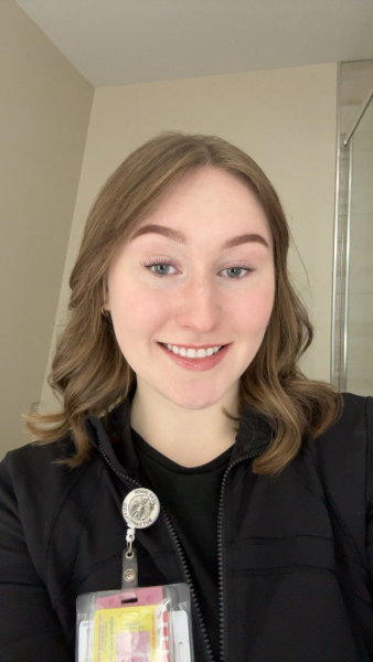 Photo of a woman with blonde hair and green eyes. She is wearing a black zip up sweater with a black shirt underneath. Her nursing badge is clipped on her sweater. 
