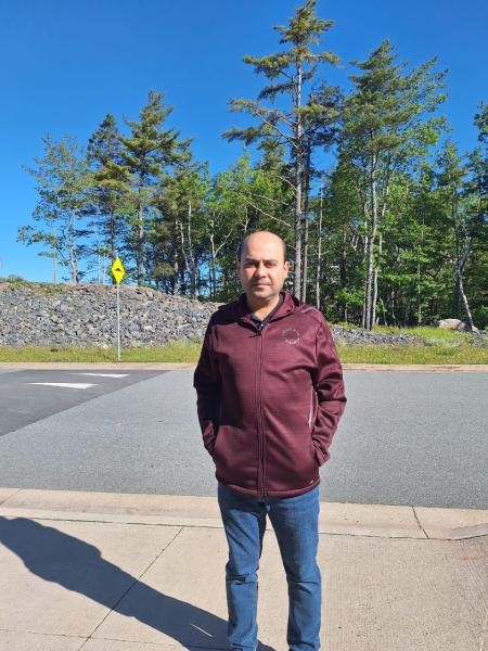 A photo of a man with short black hair. He is wearing a maroon sweater and jeans. He is standing outside with trees in the background. 