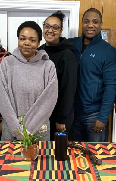 A group of people is standing in front of a table. They are smiling. The setting is indoors, with a wall in the background. The image features both men and women within the group.