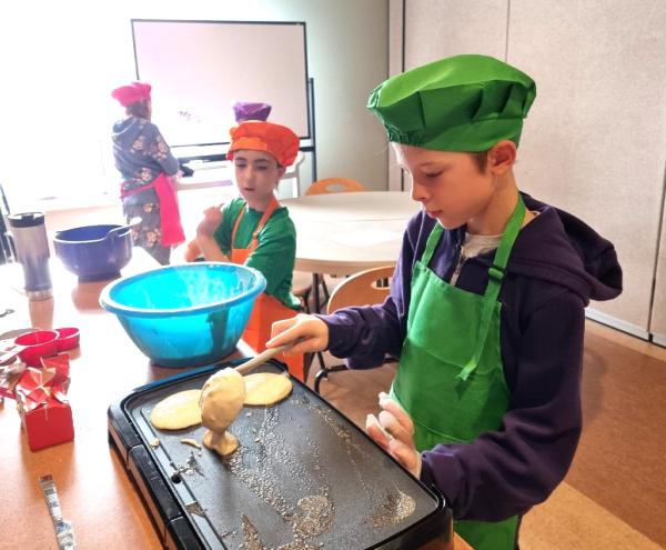 A group of kids cooking in a kitchen, wearing chef hats. They are engaged in food preparation and surrounded by various kitchen appliances and cookware.