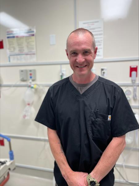 Man stands in a hospital room wearing a black scrub shirt and is smiling at the camera.