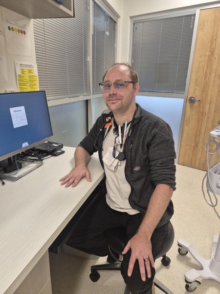 A man sits at a desk wearing a black zip-up sweater, a stethoscope, a beige shirt, and glasses and is smiling at the camera.