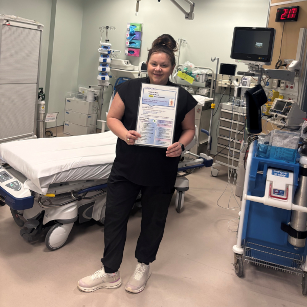 Healthcare professional standing in a hospital room holding a printed sepsis awareness document. Medical equipment and a hospital bed are visible in the background.