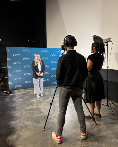 A woman stands in front of a blue backdrop with a camera person, camera and lighting person standing in front of her.