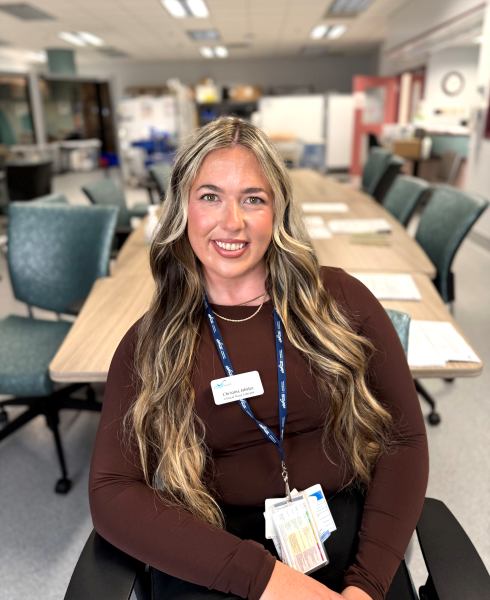 A woman with long wavy blonde and brown hair sits smiling in a meeting room. She is wearing a dark long-sleeve top, a name badge, and a blue lanyard with ID cards. The background shows a long conference table, green chairs, and hospital equipment in a bright clinical setting.