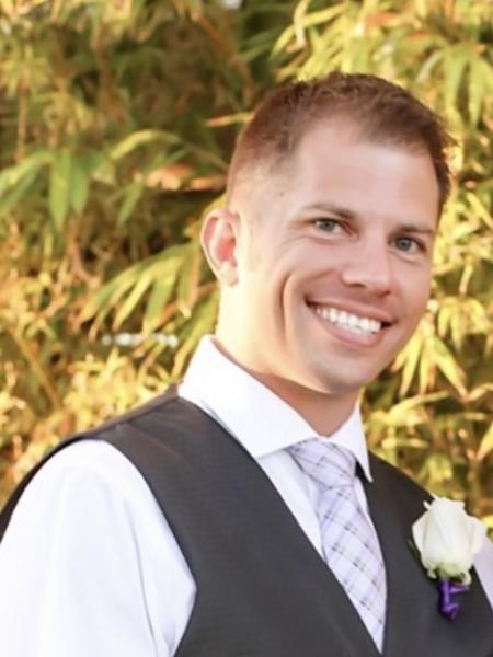 A man with short, blonde hair is wearing a vest, dress, shirt and tie is smiling at the camera.