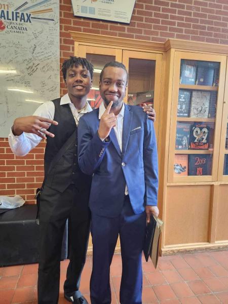 A photo of two boys. Boy on the left is wearing a black suit vest over a white button up dress shirt with black pants. Boy on the right is wearing a blue suit with white button up shirt. He is holding a diploma in his right hand.