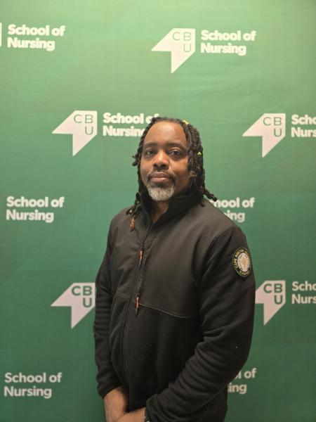 A photo of a man with brown hair and a grey goatee. He is wearing a black zip up sweater and is standing in front of a green background