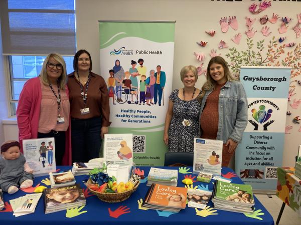 A group of four women stand side-by-side behind a table with a colourful display of promotional materials and smiling at the camera