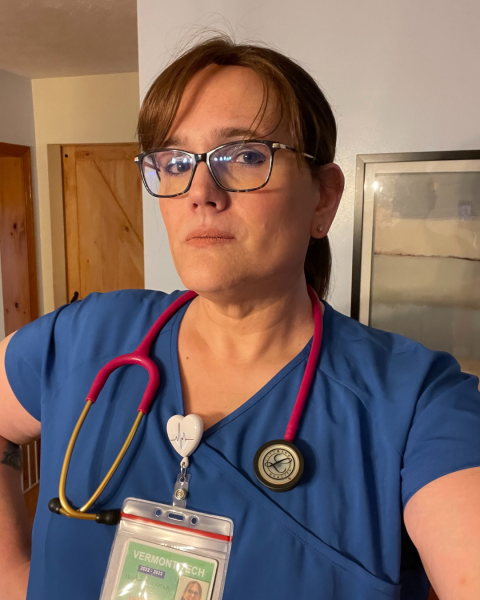 A photo of a woman with brown hair in a ponytail, she is wearing black framed glasses, blue scrubs with a red stethoscope around her neck, and a badge clipped to her collar. 