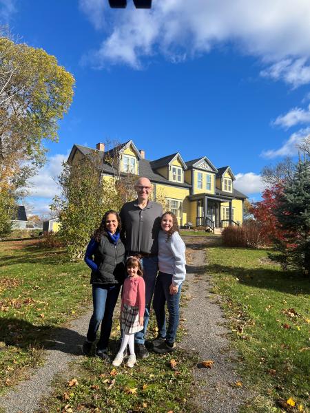 A photo of a family of four in front of a yellow home. The woman on the left is wearing a blue sweater with a black vest on top and blue jeans. She has brown curly hair. Man in the back is bald with glasses wearing a black shirt and blue jeans with a brown belt, girl on the right has curly brown hair with a light blue sweater and blue jeans. Girl in the front has brown short hair with a pink headband on, she is wearing a pink sweater with a plaid pink, black and white skirt and white tights. 