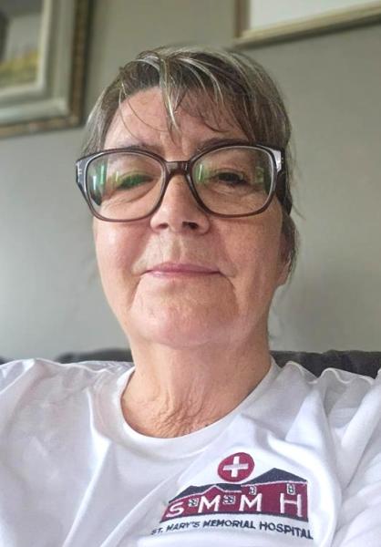 A close-up of a woman wearing glasses with a calm and gentle expression dressed in a white shirt with the logo and name "St. Mary’s Memorial Hospital" printed on it.
