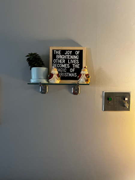 A sign sitting on a little shelf that says “The joy of brightening other lives becomes the magic of Christmas”, 2 small ornamental Santas sit in front of the sign and a small potted plant on the left side of the shelf. 