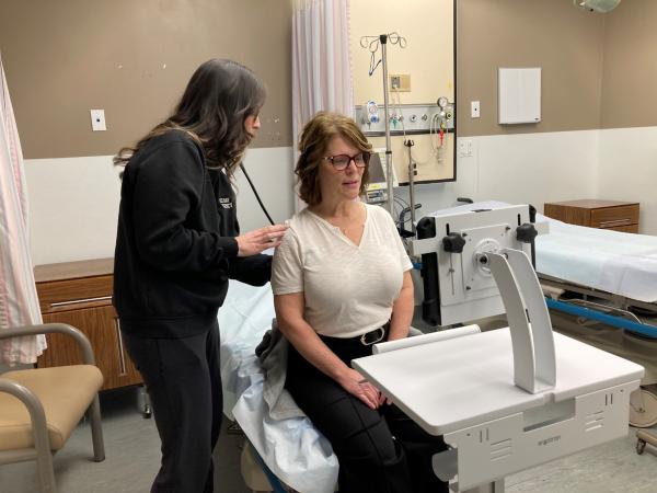 A healthcare provider assists a seated woman on a hospital examination table using a stethoscope in a clinical room.