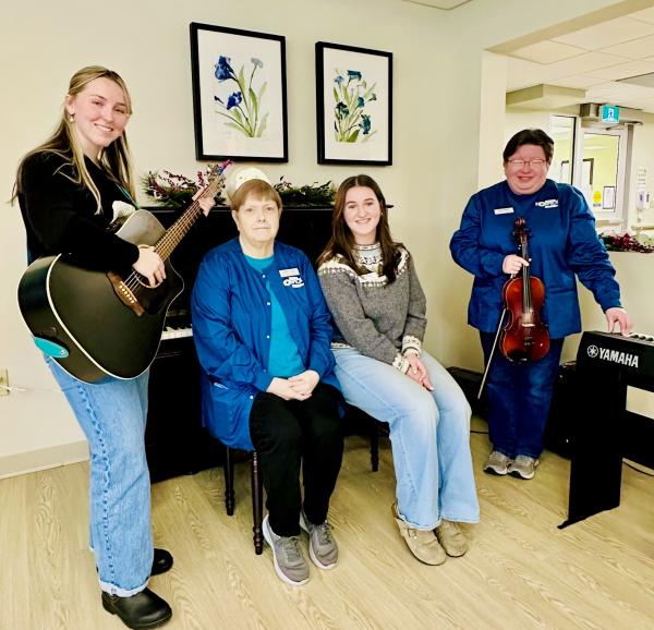 Four women pose for a photo, gather around a piano. One person holds a guitar; another person holds fiddle. The other two women sit on the piano bench. 
