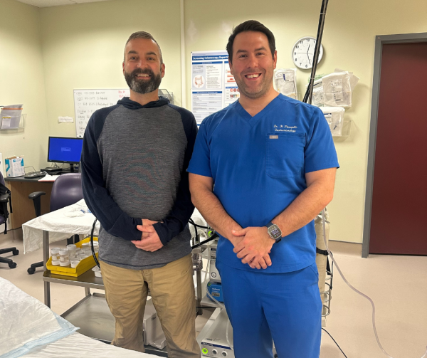 Two men standing together smiling. One in casual clothes and the other in blue scrubs. They are in a hospital room.