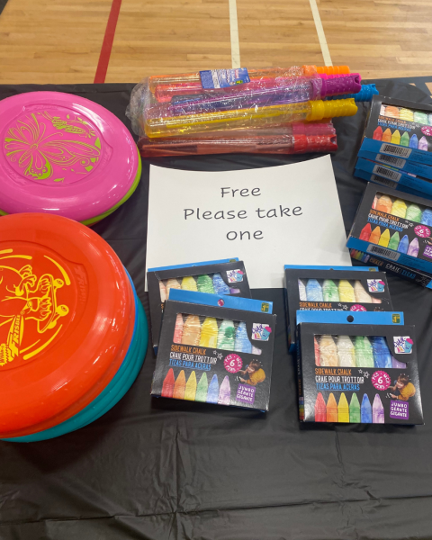 A sign reading “free please take one” displays on a table, indoors, filled with various colourful objects. 