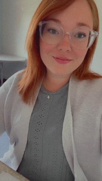  Photo of Shannon Goucher, woman with shoulder-length red hair and clear-framed glasses smiling at the camera, wearing a light grey knit top and cardigan, seated indoors.