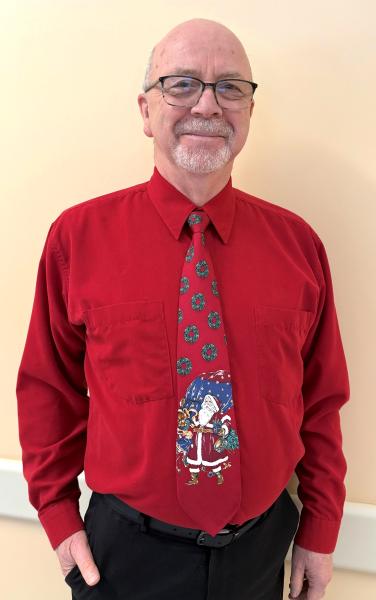 In an indoor setting, a smiling man wearing glasses is standing facing the camera. Also, he is wearing a red shirt and tie. The tie has an image of Santa Claus on it. 