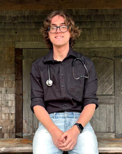 A smiling man wearing glasses, along with casual clothing - such as denim jeans and a top with sleeves - sits on an outdoor bench. A building is visible in the background. 
