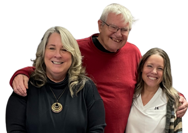 Three people smiling together indoors. On the left, a middle-aged woman with shoulder-length, blonde hair, wearing a black sweater and a gold necklace, smiles warmly. In the center, an older man with short white hair and a red sweater has his arms around both women and is smiling. On the right, a woman in her 30s with light brown shoulder-length hair, wearing a white nurse’s uniform under a striped cardigan, smiles brightly.
