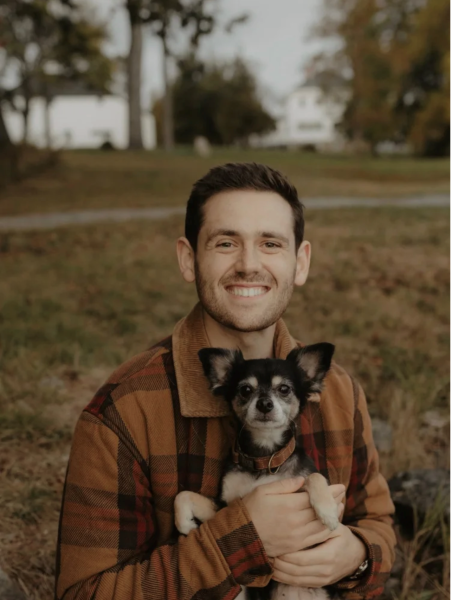 Photo of a man outside with his dog. He is wearing a brown plaid jacket and has brown hair. The dog is black and brown with a brown collar.