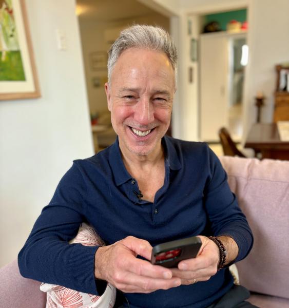 Photo of Robert Moffat siting on a couch at his residence and smiling at the camera while using the YourHealthNS app on his phone.