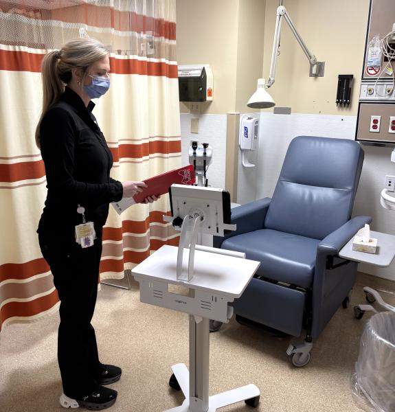 A masked healthcare worker holding a red clipboard stands beside an iPad on a workstation in a hospital exam room.