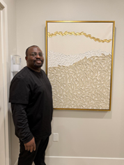 Dr. Adebayo stands in a clinic hallway beside a framed abstract artwork in neutral tones with gold accents, wearing black clinical attire and smiling toward the camera.