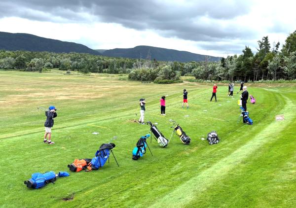 A group of people gathered on a golf course. The scene features lush grasslands under a clear sky with clouds and trees in the background. 