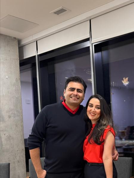 A photo of a man and a woman. The man is on the left and has brown hair. He is wearing a black sweater over a red collared shirt. The woman is on the right and she has long brown hair. She is wearing a red blouse with black pants.