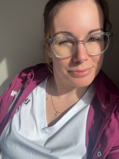 A woman sitting by a window. She has white scrubs on with a pink sweater and a stethoscope around her neck. She is wearing clear framed glasses. Her hair is brown and is tied back.