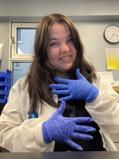A woman sitting by a window. She has white scrubs on with a pink sweater and a stethoscope around her neck. She is wearing clear framed glasses. Her hair is brown and is tied back.