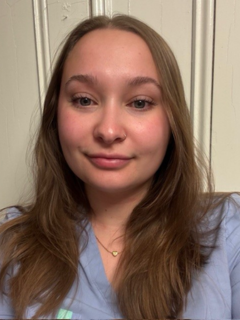 A woman with long light brown hair. She is wearing blue scrubs.
