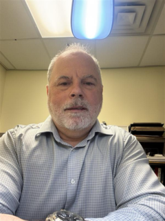A man in a office with a blue buttoned shirt. He has white hair with a white beard.