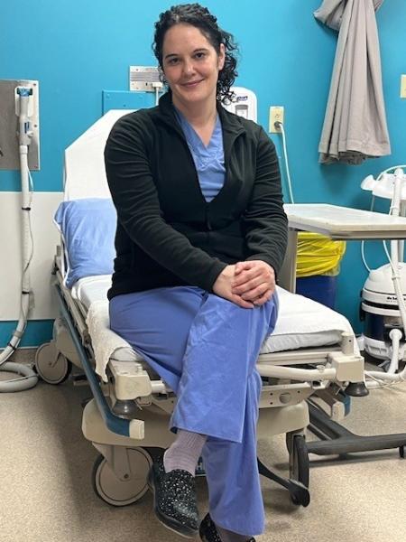 A woman is wearing blue scrubs with a black sweater and is sitting on a hospital bed, smiling at the camera.