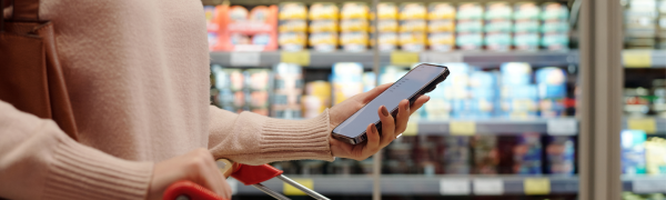 You see the torso of a woman in a grocery store pushing a cart with her cell phone in her hand.