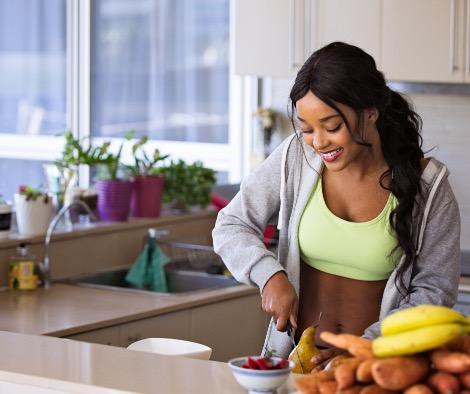 Black woman in a kitchen preparing a healthy meal. She is wearing a zippered grey hoodie and a green sports bra.