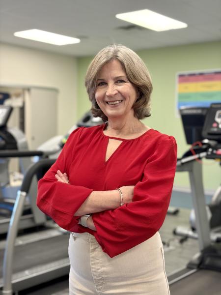 A woman stands in a gym wearing a red shirt and beige pants with her arms folded, smiling at the camera.